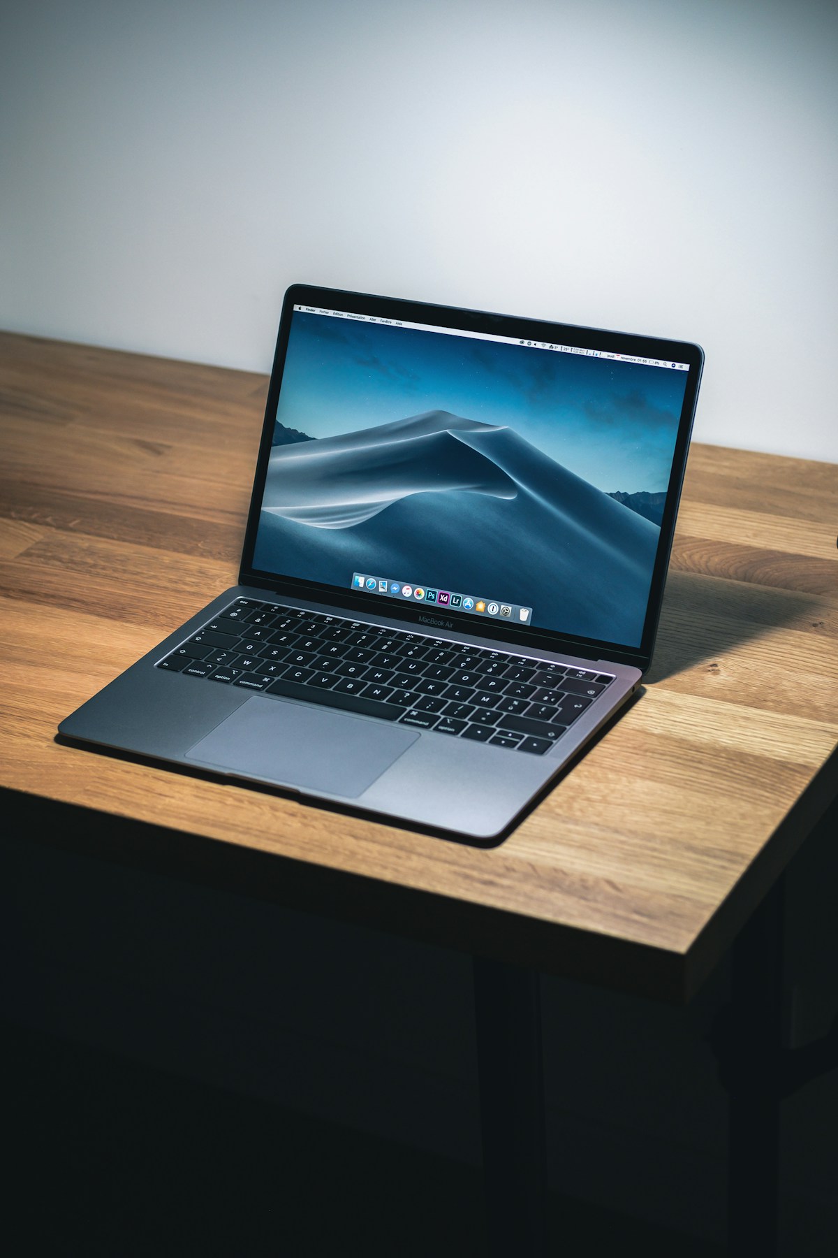 An open MacBook on a simple wooden desk, lit by natural light.